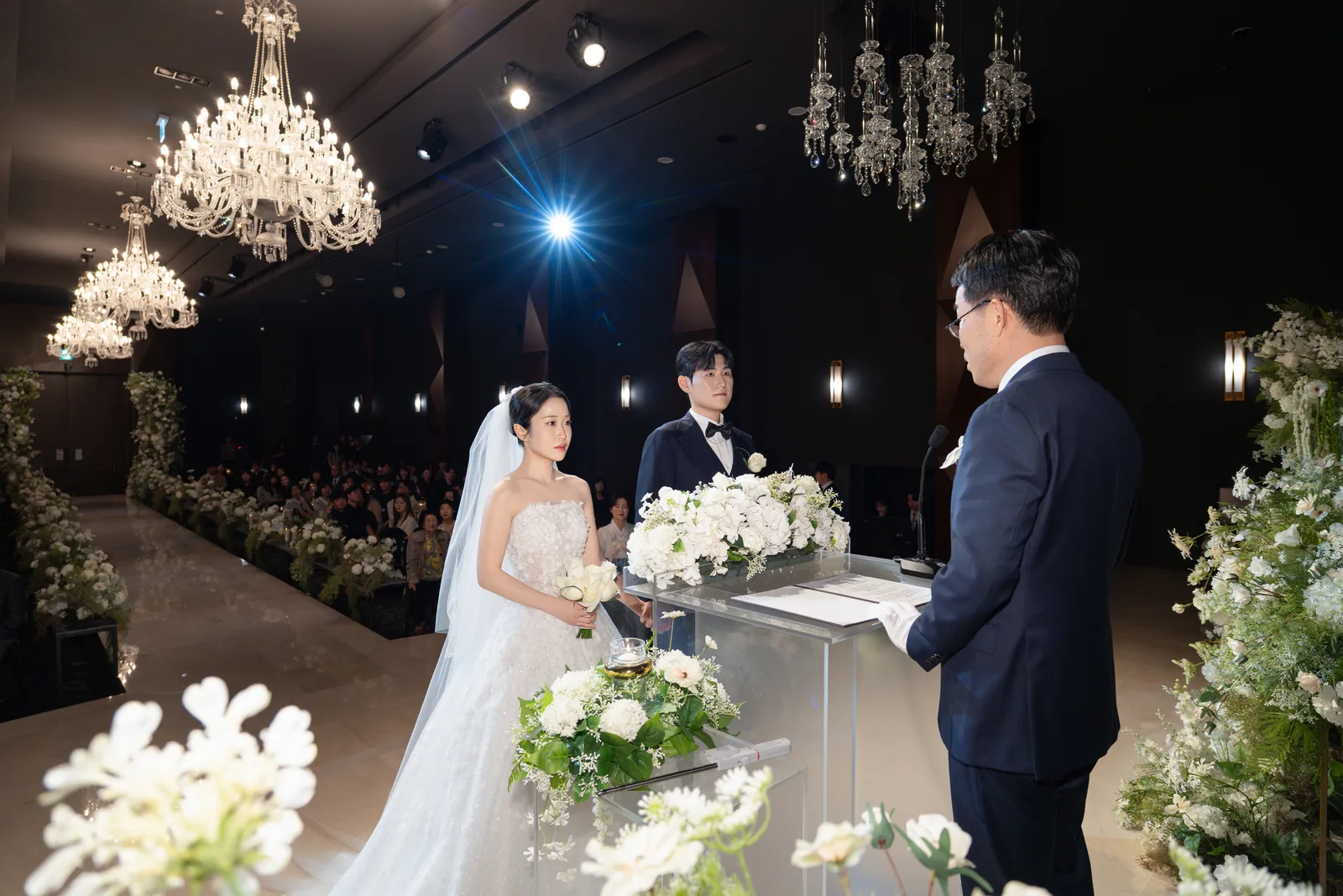 크리스털 샹들리에 아래 흰 꽃이 놓인 예식대 건너편 정장 차림 진행자를 바라보는 신랑 신부