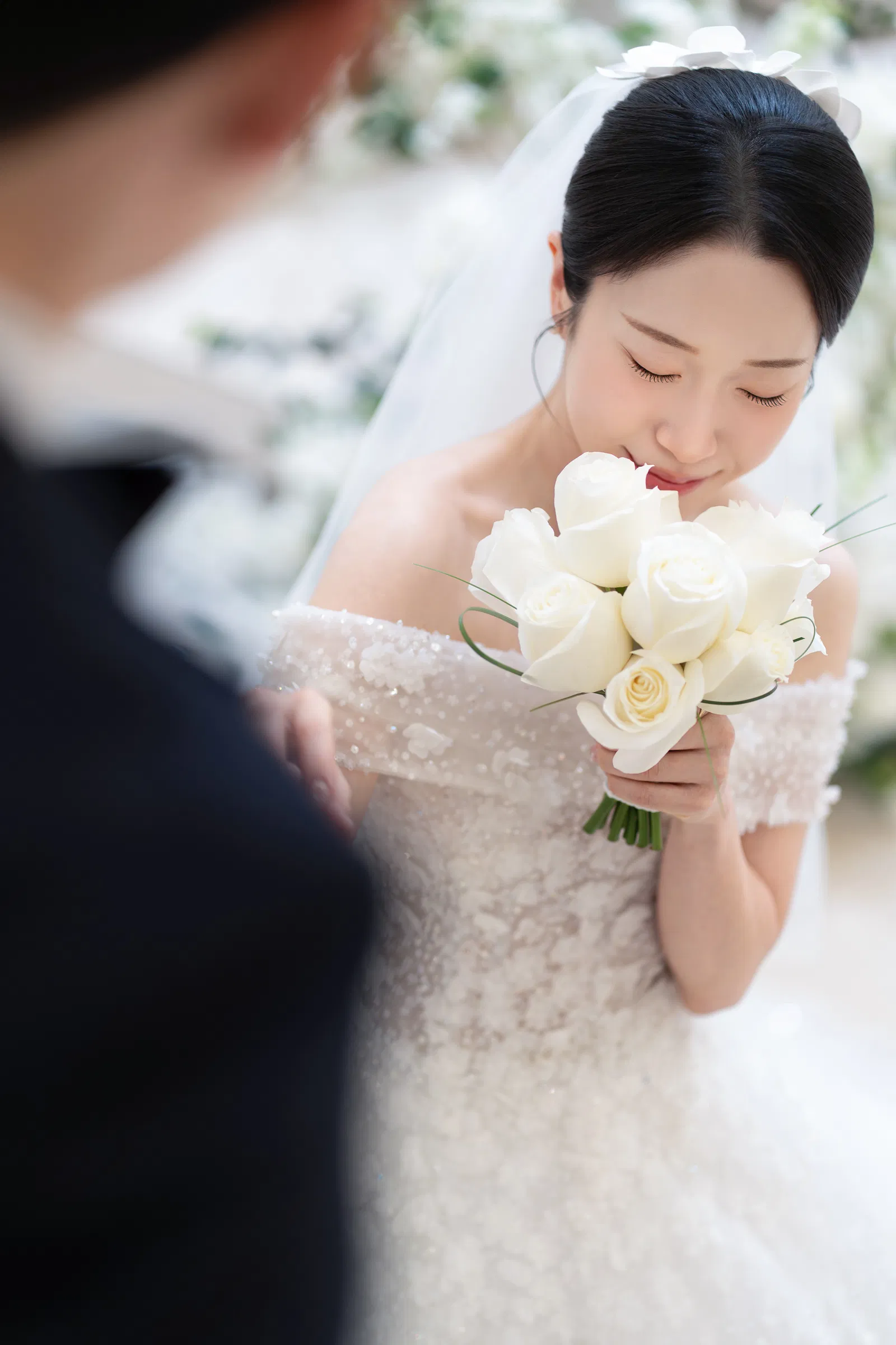 흰 장미 부케를 내려다보는 신부와 흐릿하게 보이는 신랑의 옆모습