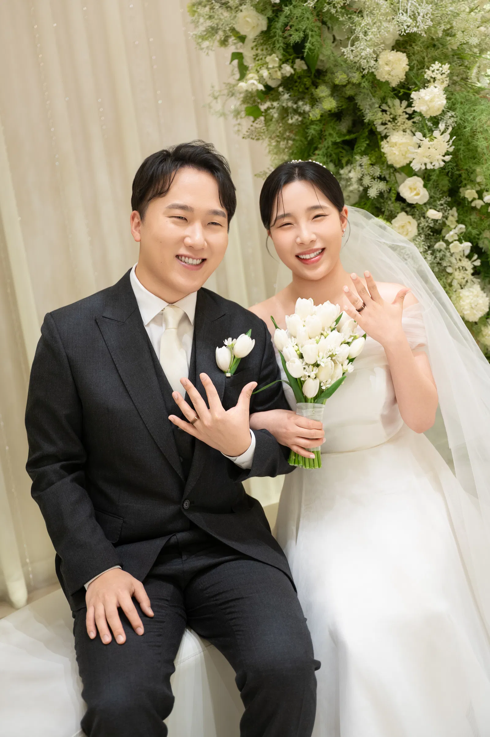 꽃 장식 앞에서 신랑은 앉고 신부는 부케를 든 채 두 사람 모두 한 손을 앞으로 보인 사진