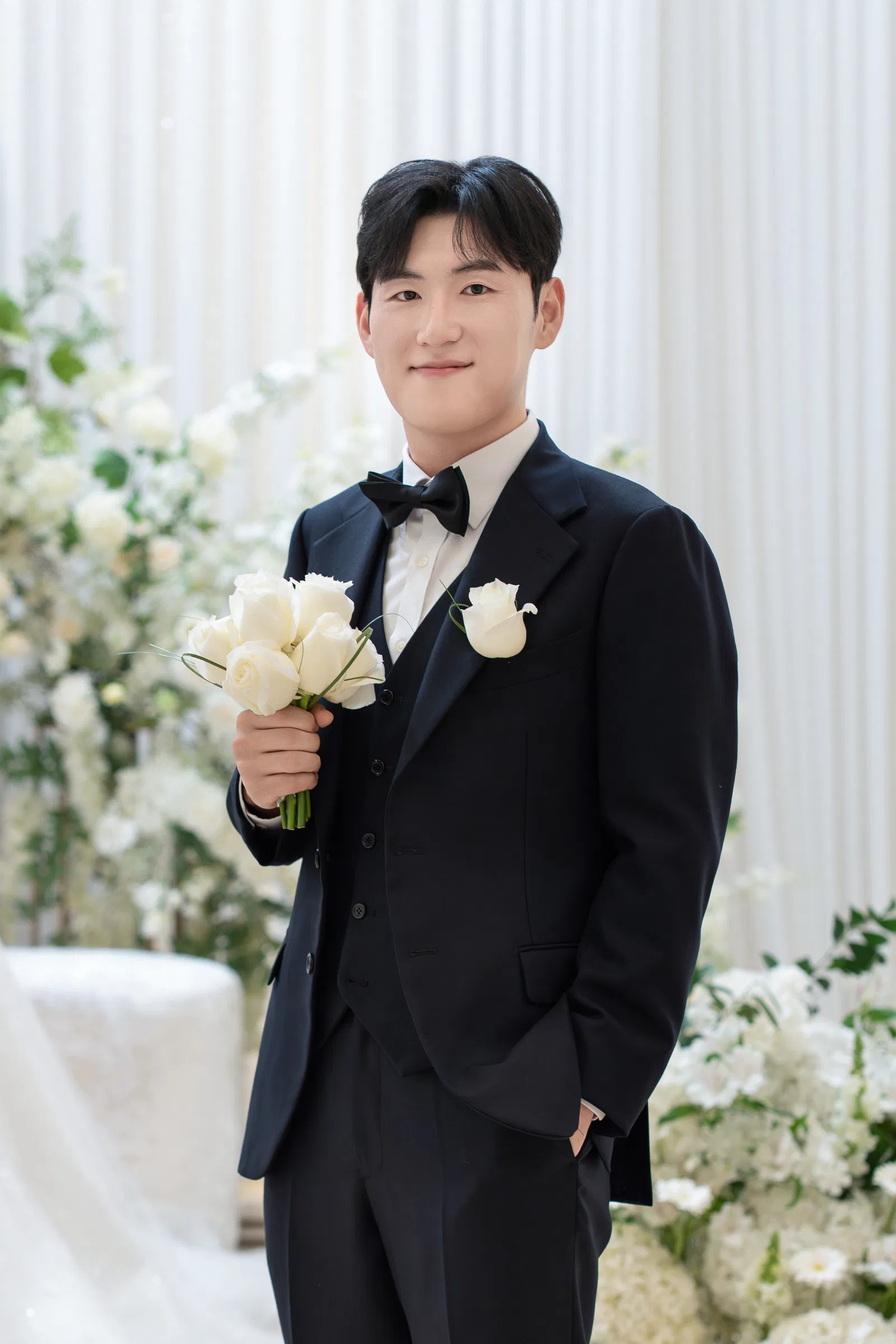 연한 꽃과 커튼 앞에서 작은 흰 장미 부케를 들고 홀로 선 검은 턱시도 차림 신랑