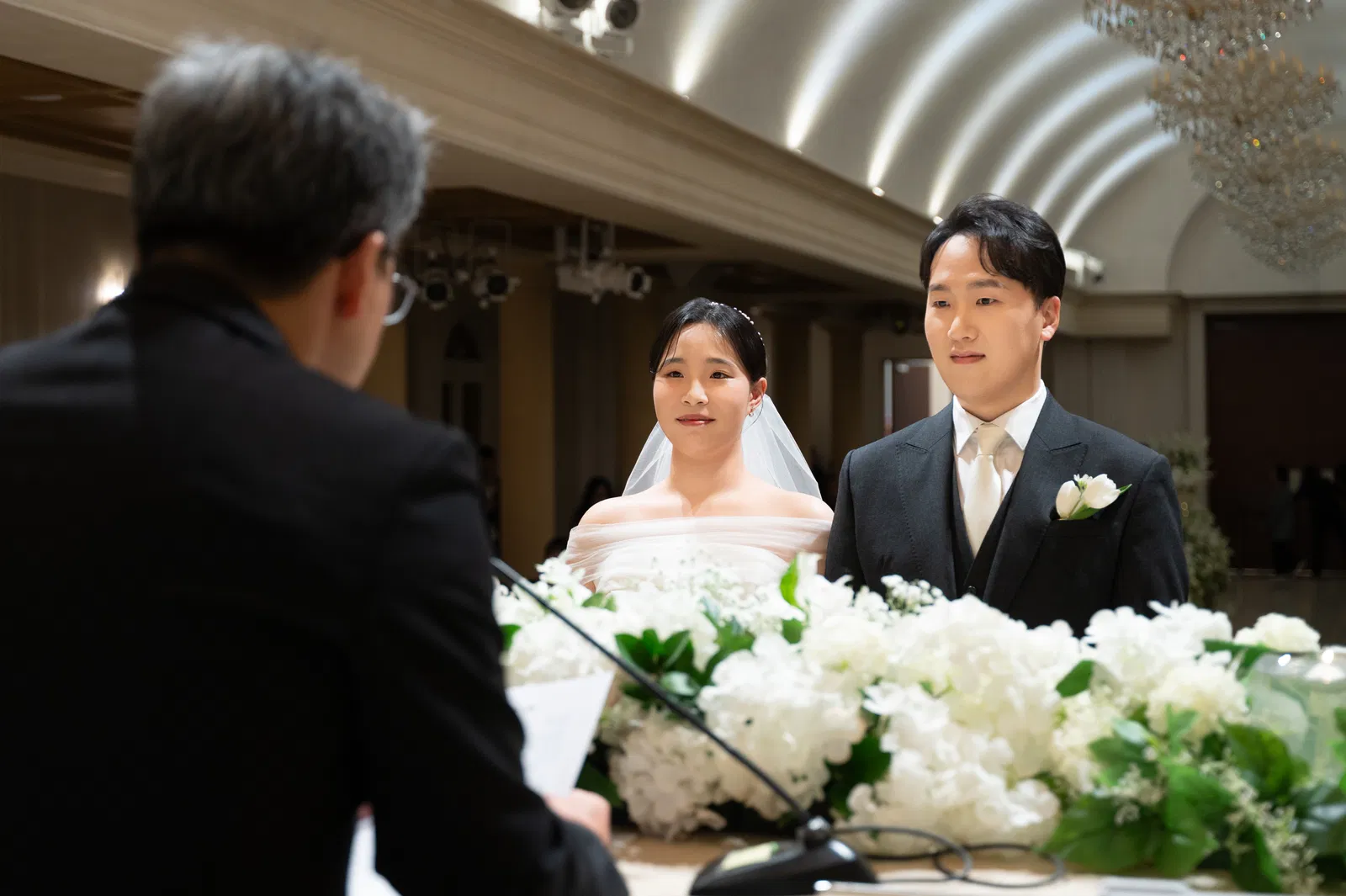 흰 꽃이 놓인 예식대 건너편 정장 차림 진행자를 바라보는 신랑 신부