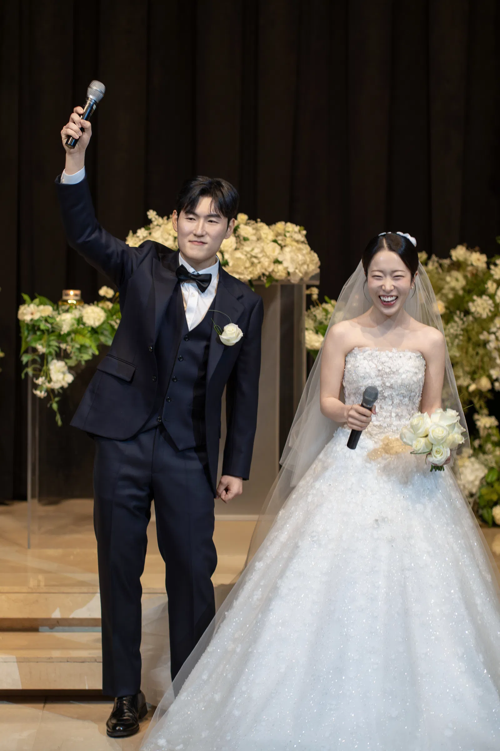예식 단상에서 신랑은 마이크를 머리 위로 들고 신부는 마이크와 흰 부케를 든 모습