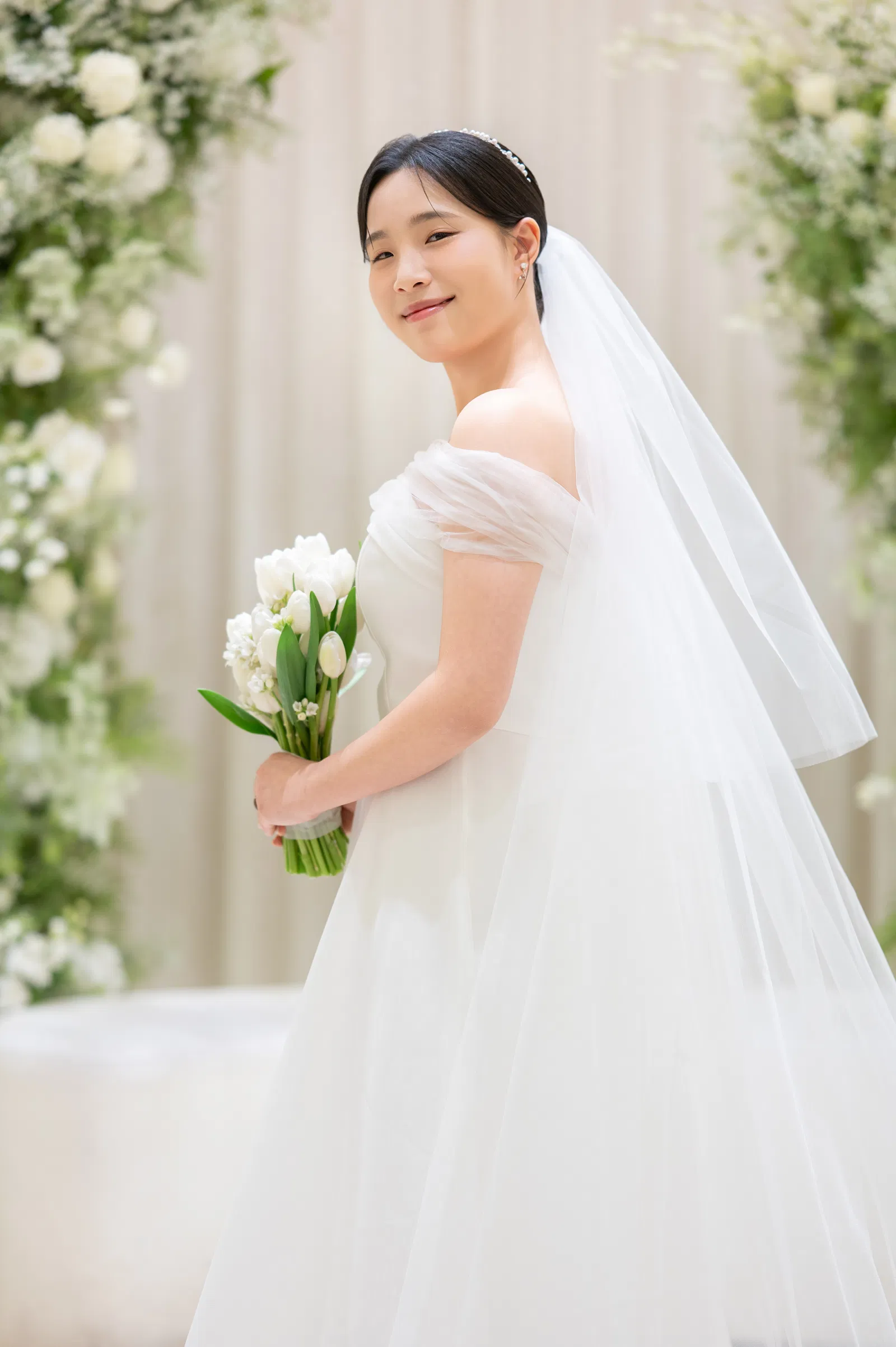 흰 꽃 사이에서 튤립 부케를 들고 뒤를 돌아보는 오프숄더 드레스 차림 신부