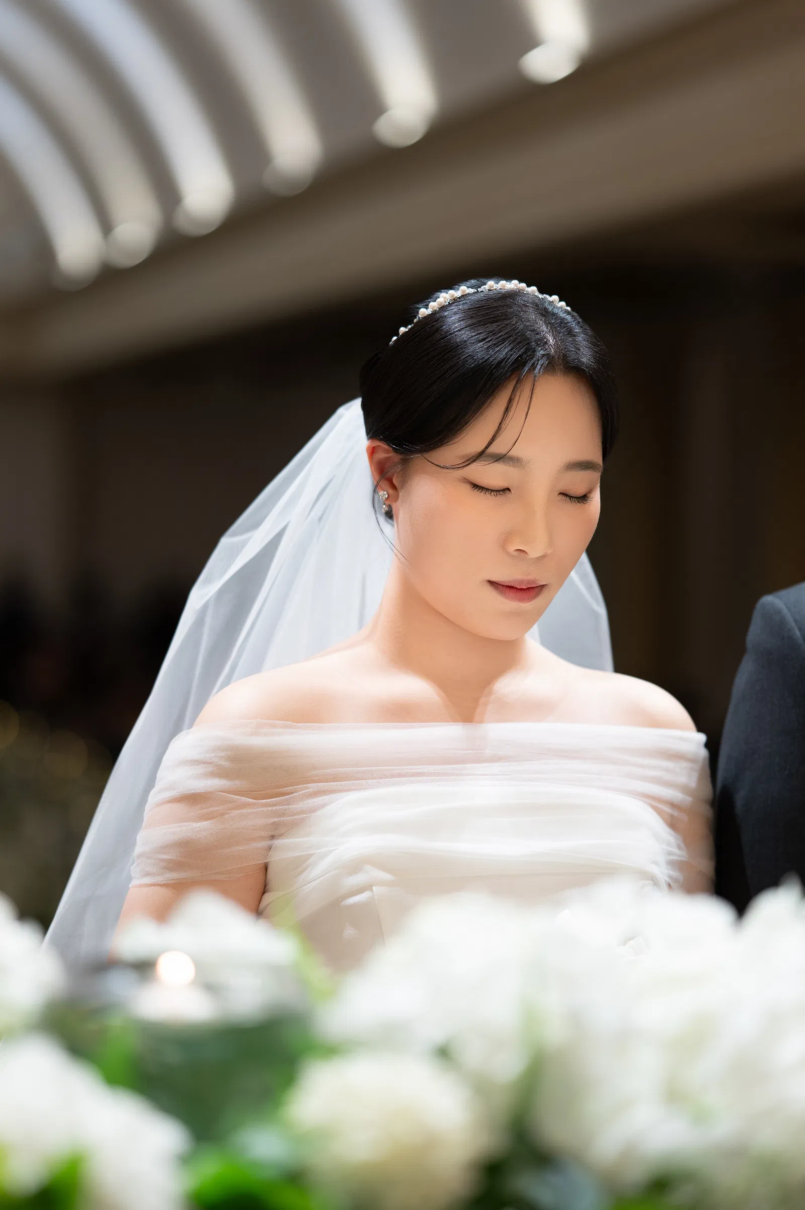 곡선형 천장 조명 아래 흰 꽃 너머에서 아래를 바라보는 신부의 상반신