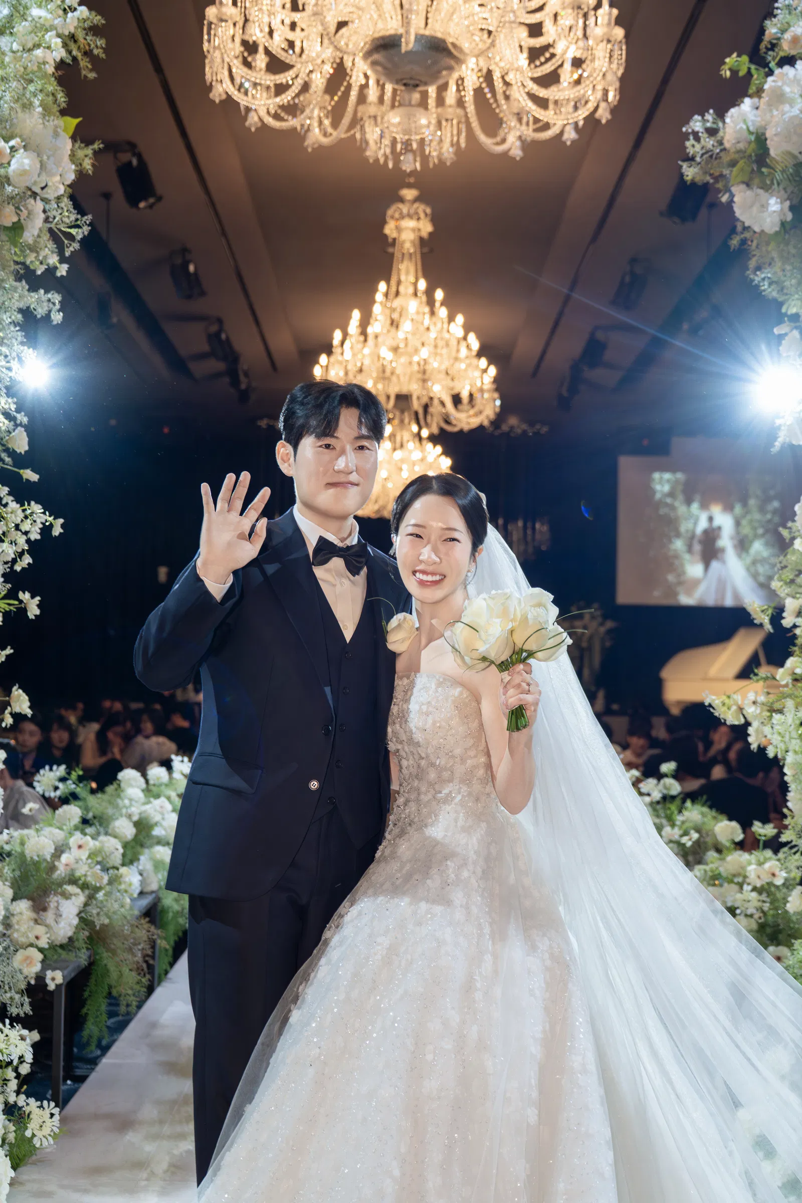 샹들리에 아래 나란히 한 손을 들어 보이는 신랑 신부와 오른쪽으로 길게 이어진 베일