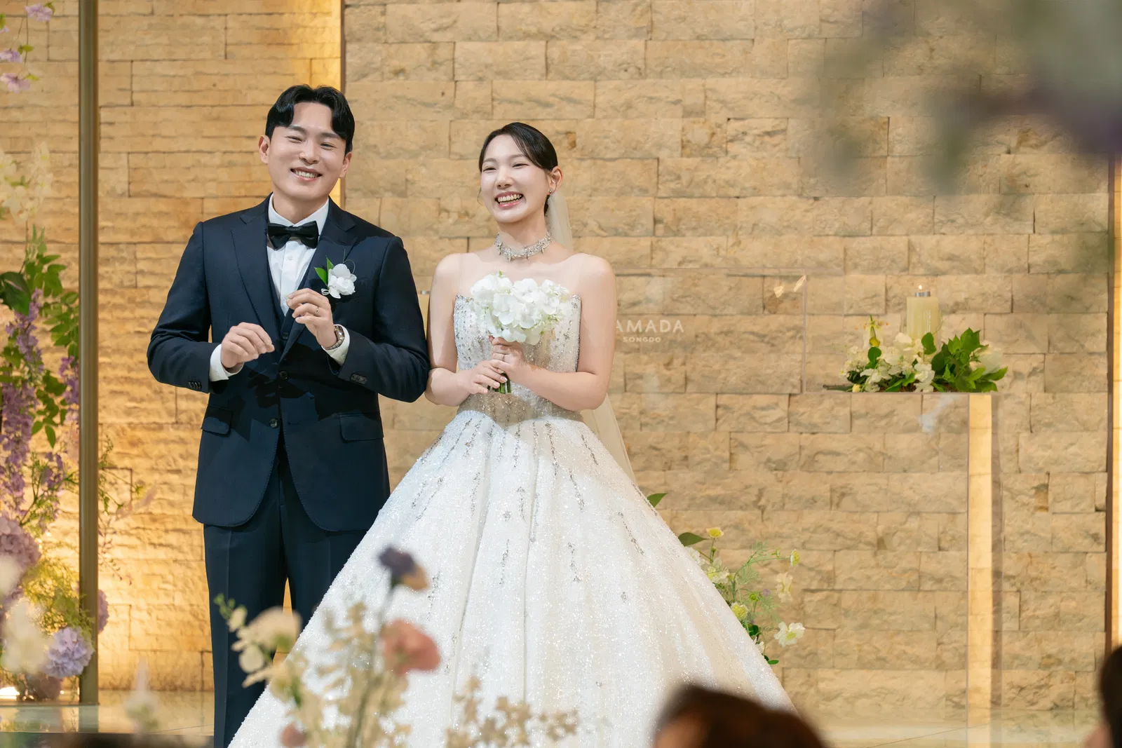 라마다호텔 송도점 따뜻한 벽면과 꽃장식 앞에서 웃으며 나란히 선 신랑과 신부